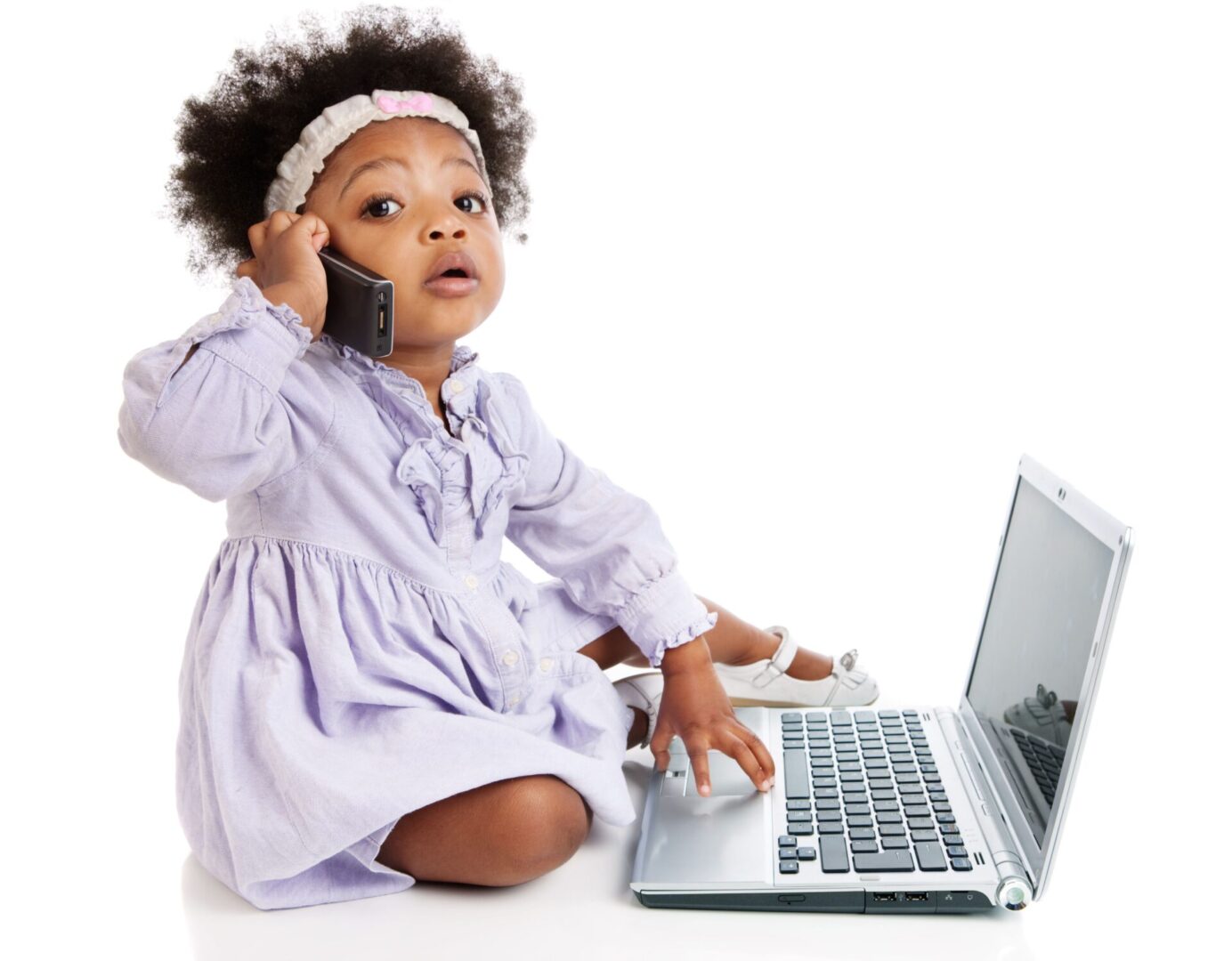 A baby girl sitting on the floor next to a laptop.